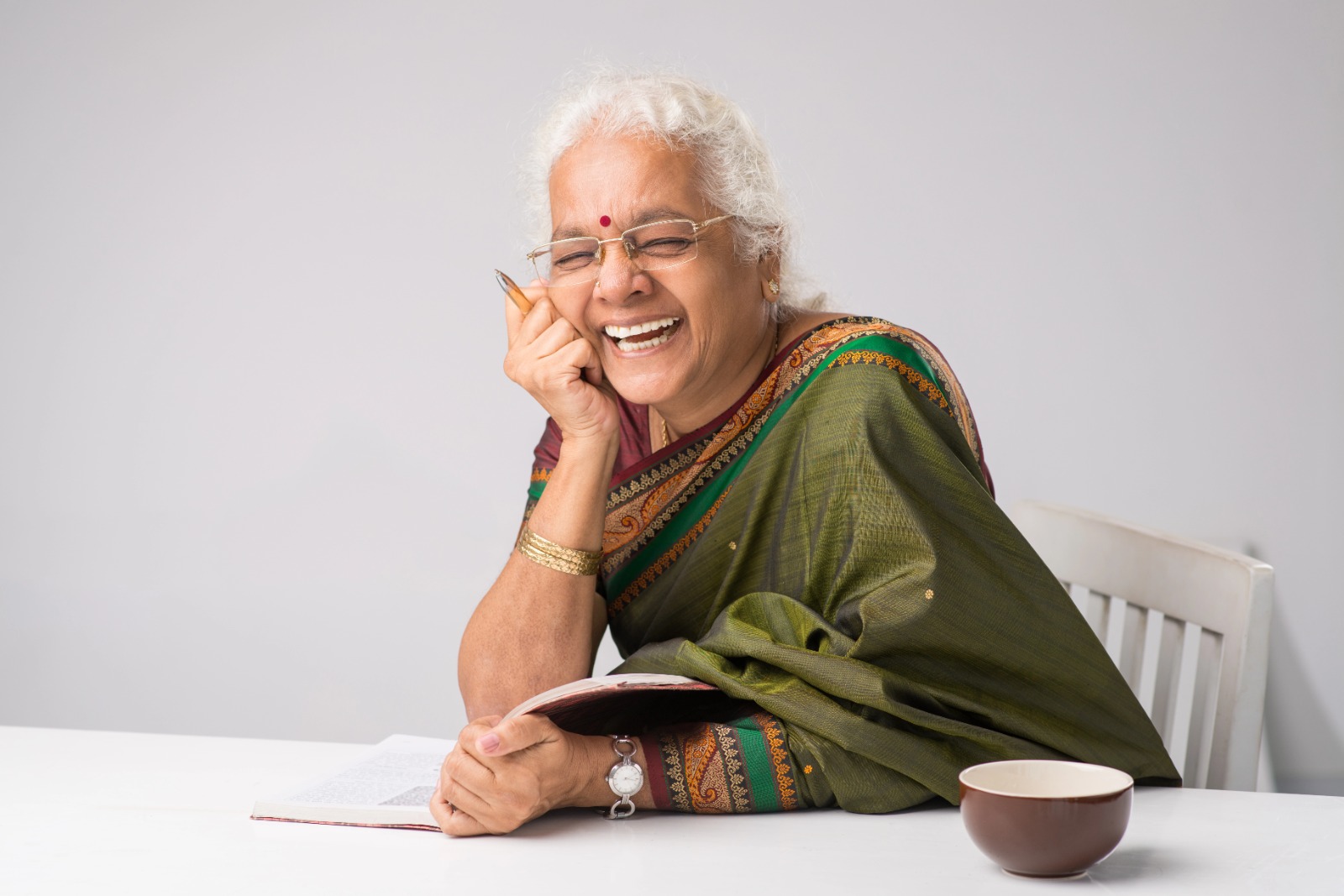 Elderly Indian woman in green saree smiling joyfully while reading a book, sitting at a white table with a cup beside her.