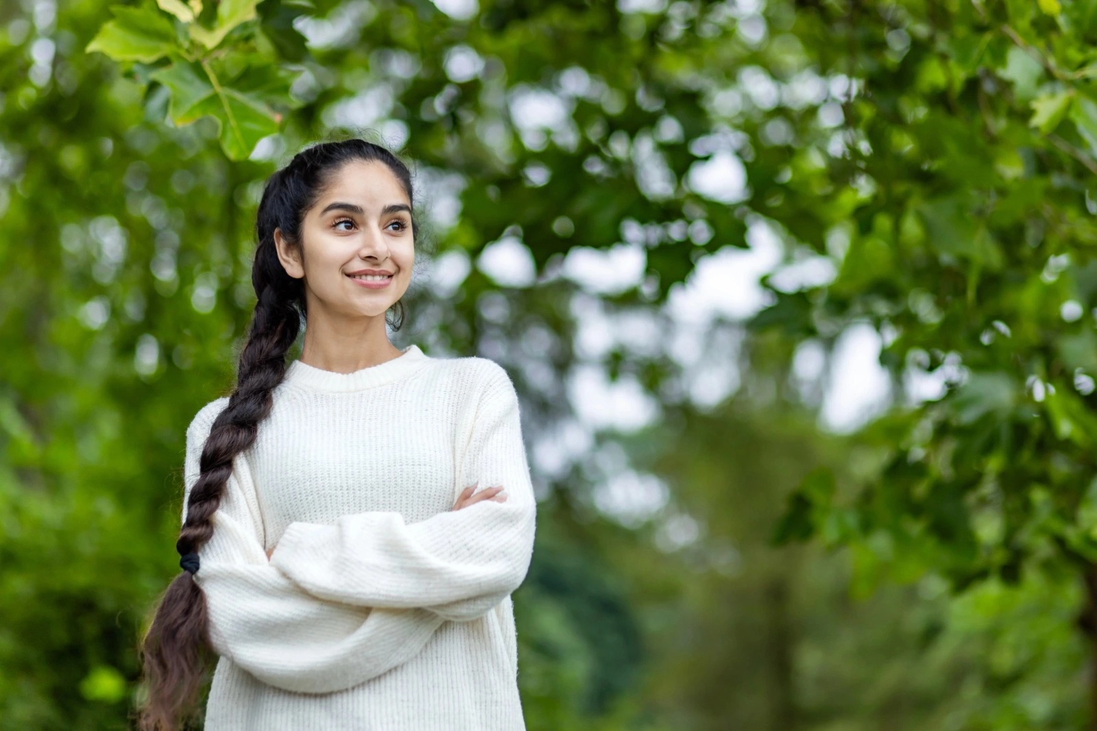 A happy young Indian woman with long braided hair wearing a cozy white sweater, smiling confidently outdoors with lush green trees in the background, symbolizing health and positivity