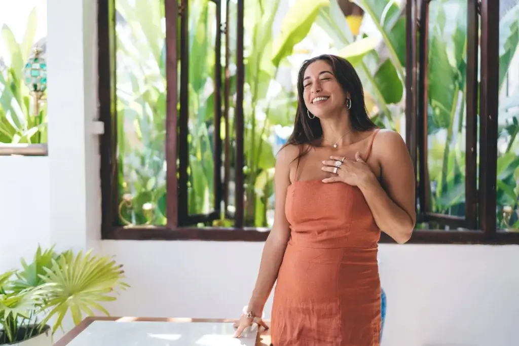 A South Asian woman in a rust dress smiling with serenity, hand on her heart, in a sunlit tropical space—radiating balance and confidence, representing the healing power of Miror PCOS.