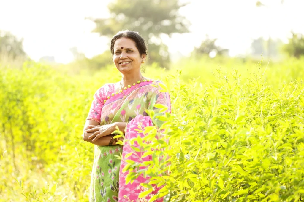 Smiling Indian woman in a pink and green saree standing in a lush green field, radiating post-menopause confidence with Miror Thrive support.