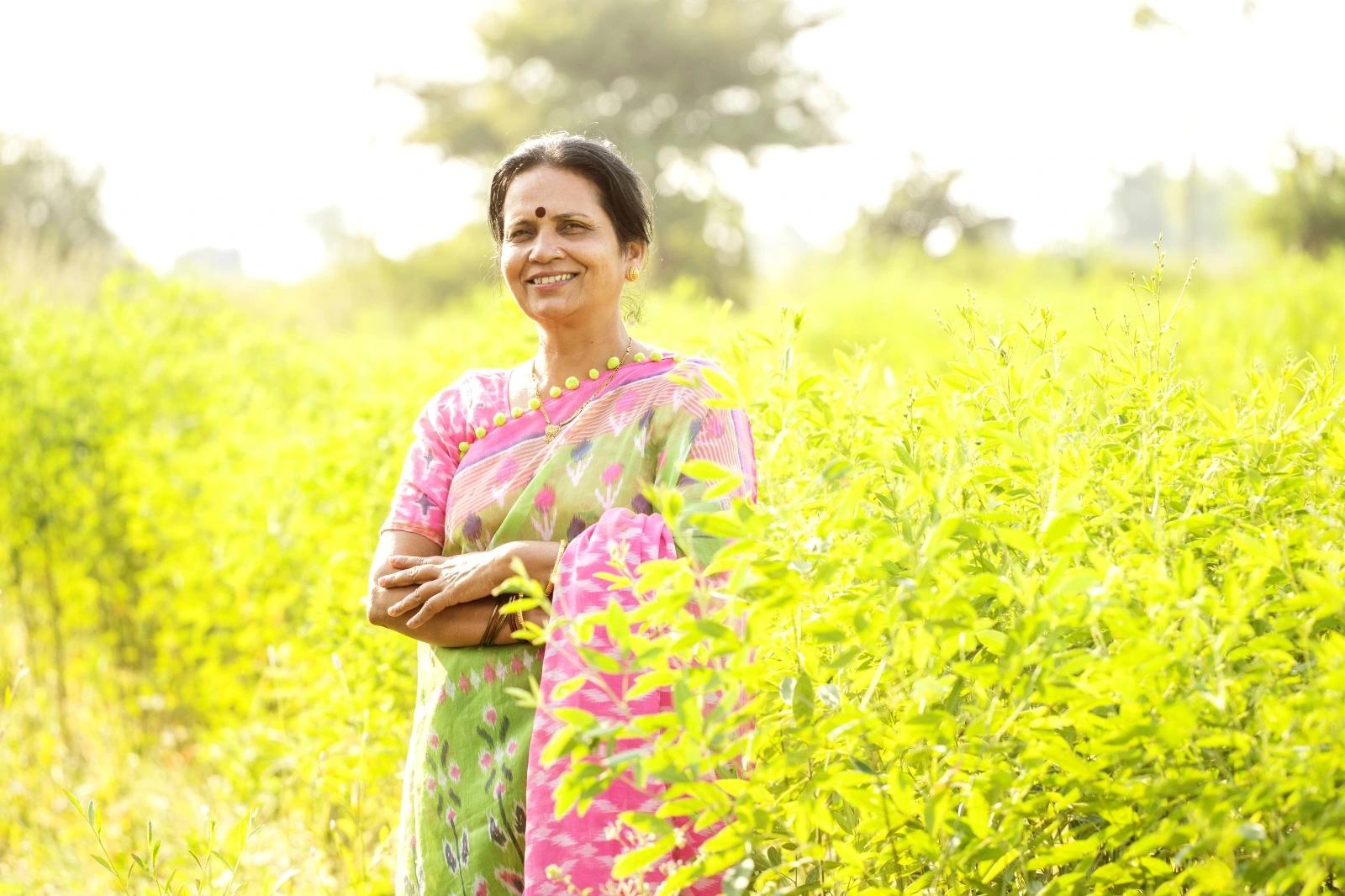 Smiling Indian woman in a pink and green saree standing in a lush green field, radiating post-menopause confidence with Miror Thrive support.