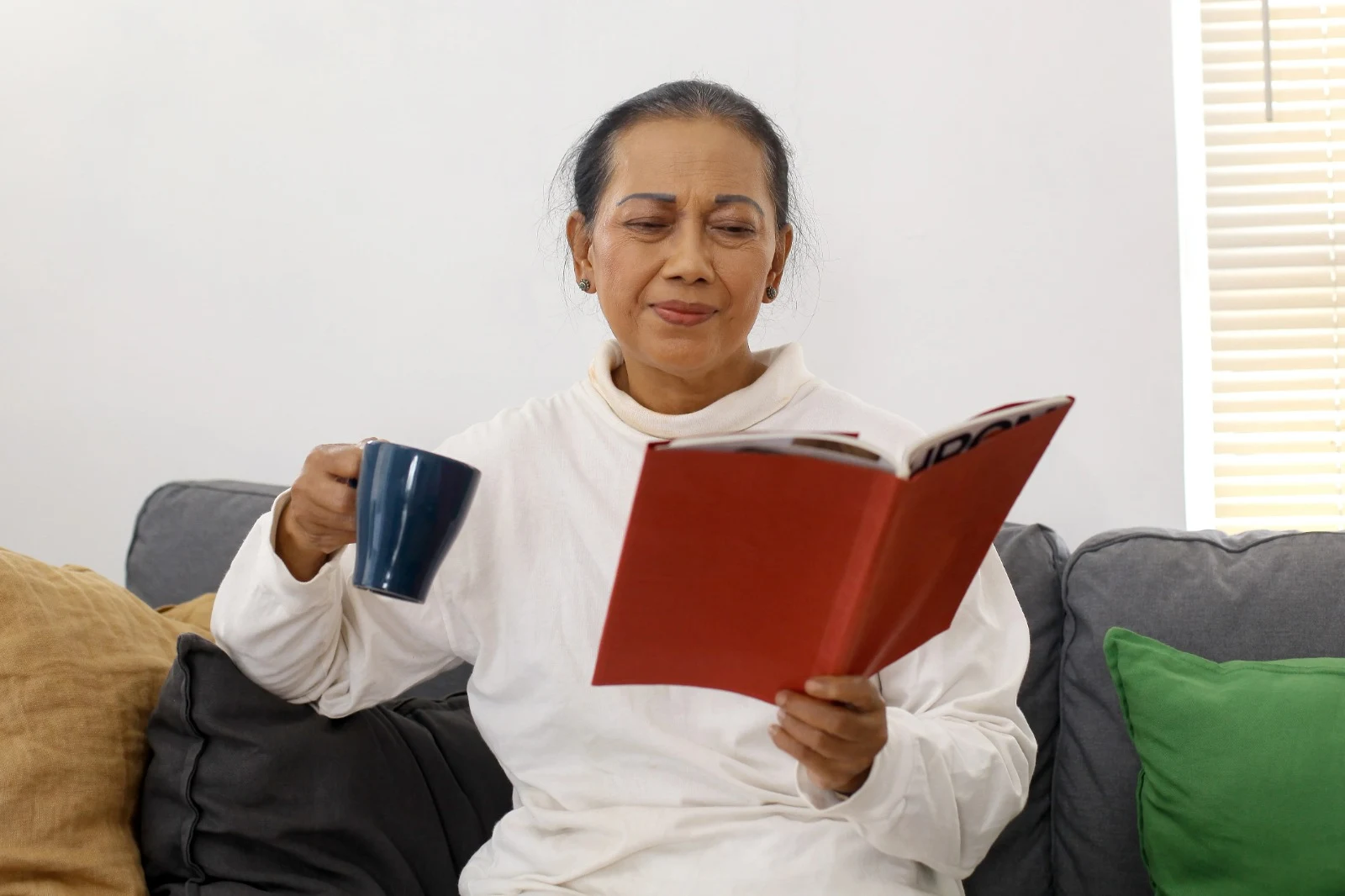 Senior Indian woman sitting on a couch reading a book while holding a blue mug, wearing a white sweater, surrounded by colourful cushions.