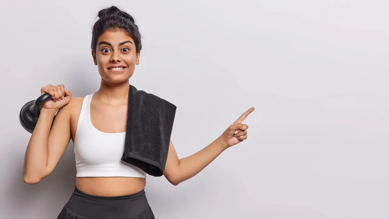 A smiling young woman in workout clothes holding a kettlebell and pointing sideways with a towel on her shoulder.