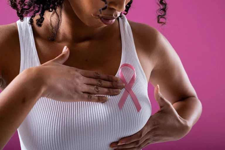 A woman wearing a white tank top performs a breast self-exam while wearing a pink ribbon — a global symbol of breast cancer awareness — against a pink background.