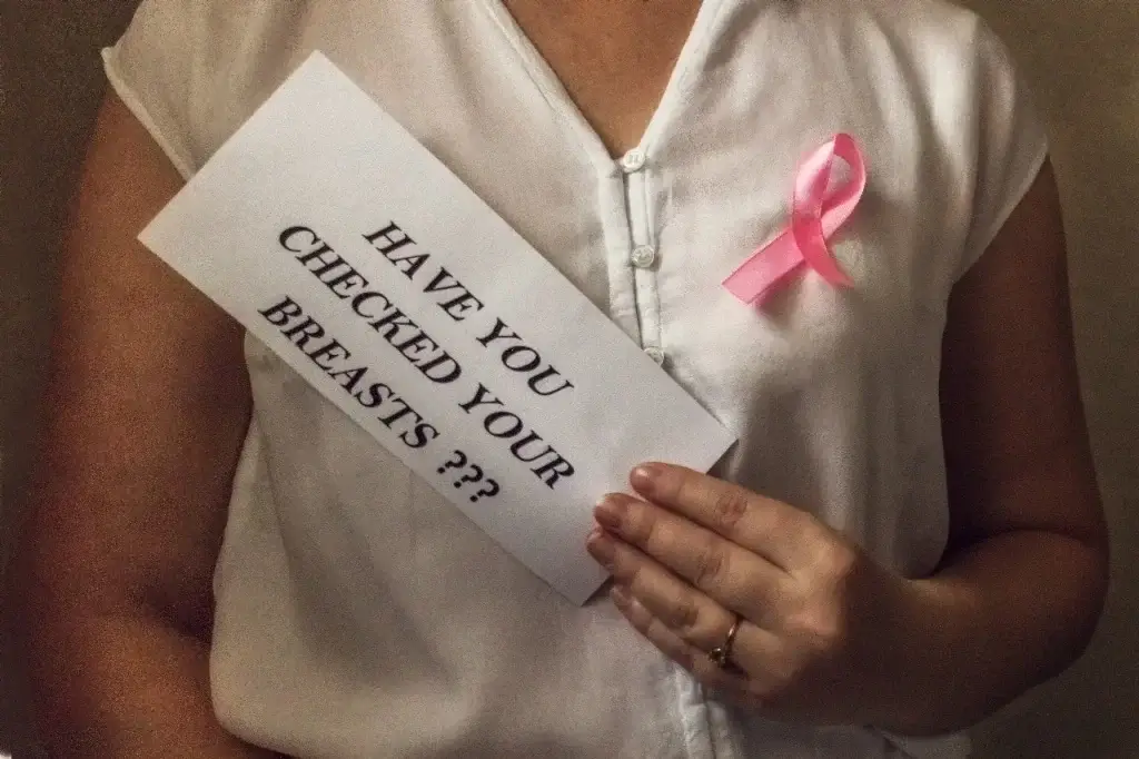 A woman wearing a white shirt with a pink ribbon holding a sign that says “Have you checked your breasts?” promoting breast cancer awareness and self-exam.