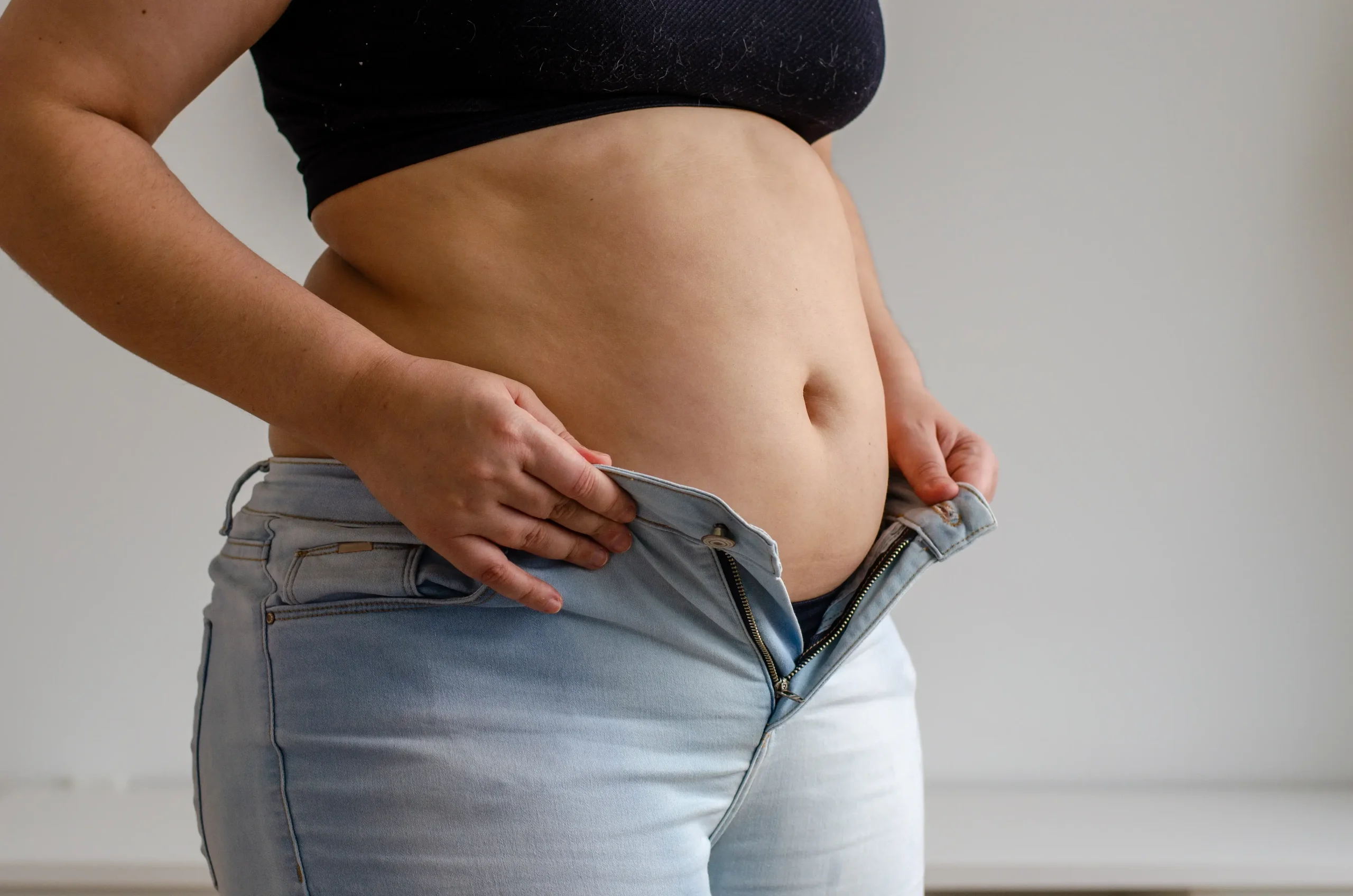 A woman with a visible midsection trying to zip her jeans, showing common abdominal changes many women experience after 40.