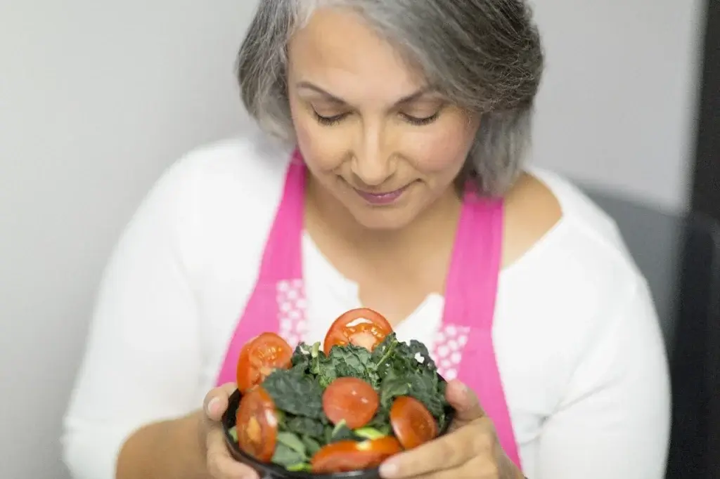 Woman with gray hair in a pink apron holding a bowl of fresh kale and tomatoes, looking down gently at the meal.