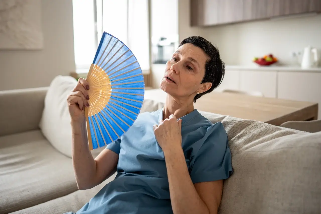 A middle-aged woman experiencing a hot flash, sitting on a couch and cooling herself with a handheld fan.