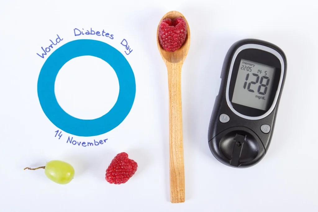 Blue circle symbol for World Diabetes Day with date 14 November, alongside a glucose meter reading 128 mg/dL, raspberries, grape, and a wooden spoon on a white background.