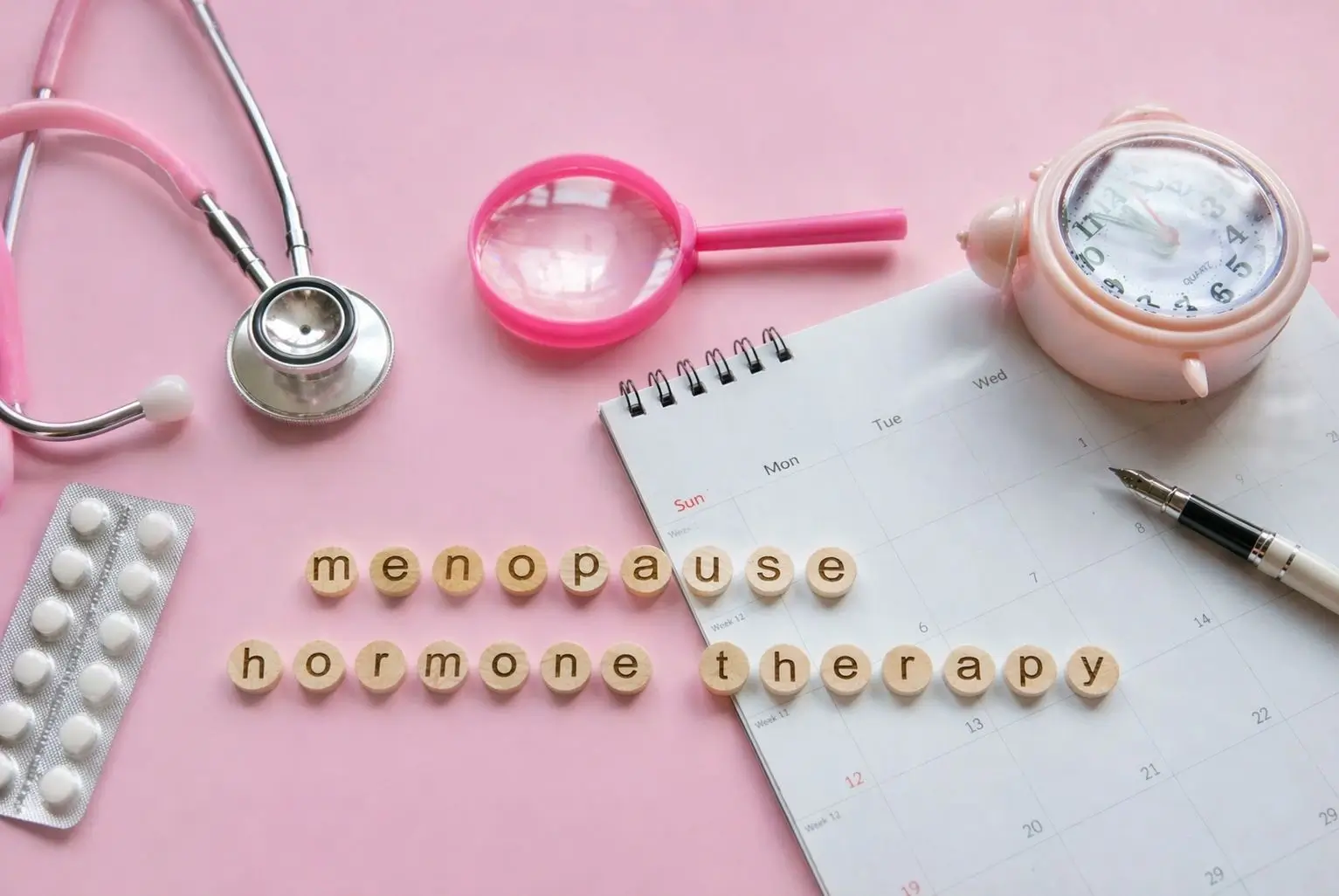 Menopause concept flat lay with wooden letters spelling menopause and hormone therapy, stethoscope, pills, calendar, and clock on a soft pink background